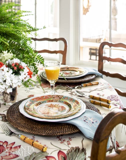 Dining table set with floral tablecloth, woven placemats, ornate plates, bamboo-handled cutlery, and a glass of orange juice.