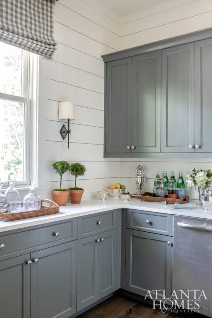 Gray kitchen cabinets with white shiplap walls, a window with a gingham shade, and a white quartz countertop with barware and decorative plants.