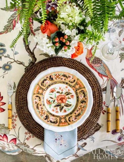 Dining table set with a floral patterned tablecloth, woven placemat, white charger plate, and a decorative plate with coral and navy floral design.