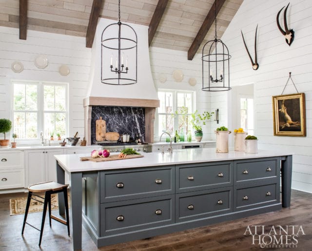 Farmhouse kitchen with white shiplap walls, vaulted wood-beamed ceiling, and a large gray island with a white countertop. Features include a dark marble backsplash, cage pendant lights, and mounted animal horns.