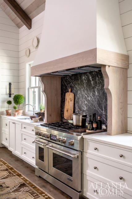 White kitchen with white shiplap walls, a large wooden range hood with carved details, a black marble backsplash, and stainless steel appliances.