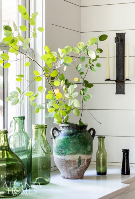 Green glass bottles and a large distressed green ceramic vase with branches sit on a white countertop. A black sconce with two cream candles hangs on a shiplap wall.