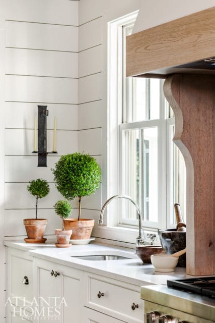 White kitchen with shiplap walls, a sink beneath a window, and decorative topiary plants in terra cotta pots.