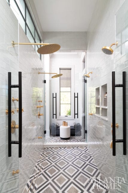 Modern bathroom with multiple gold shower heads, glass shower enclosures, black hardware, and geometric floor tiles.