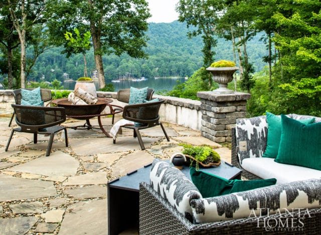 Stone patio with fire pit, wicker chairs, and sectional sofa overlooking a lake surrounded by trees.
