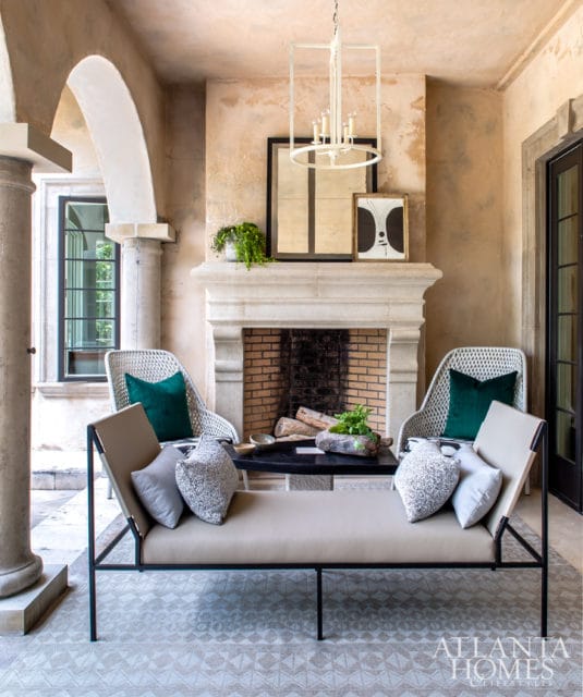 Covered patio with a stone fireplace, white wicker chairs with green pillows, and a tan chaise lounge with decorative pillows.