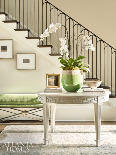 Entryway with a white console table topped with orchids and books, a green velvet bench, and framed art on the wall next to a staircase with black railings.