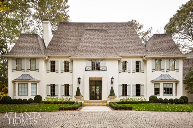 White stucco French Provincial style home with a mansard roof, light blue shutters, and a cobblestone driveway.
