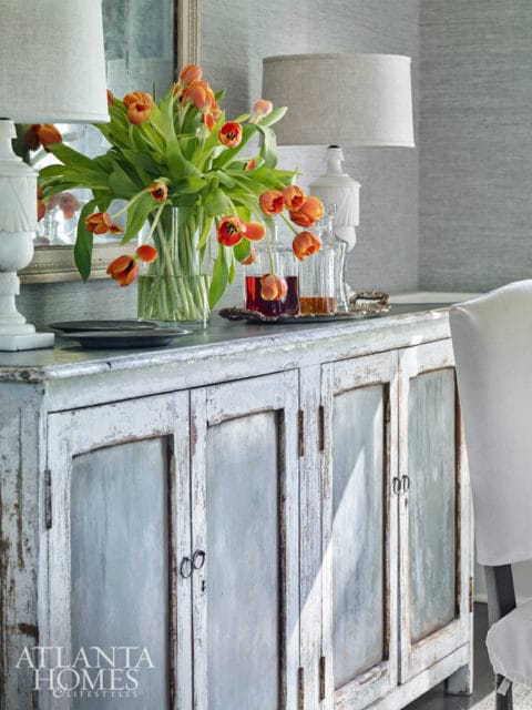 Distressed antique cabinet with glass doors, topped with a vase of orange tulips, decanters of amber liquid, and lamps against a textured grey wall.