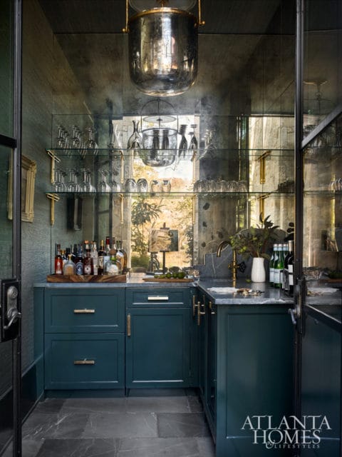 Dark teal bar cabinet with brass hardware, marble countertop, glass shelves with glassware, and mirrored walls.