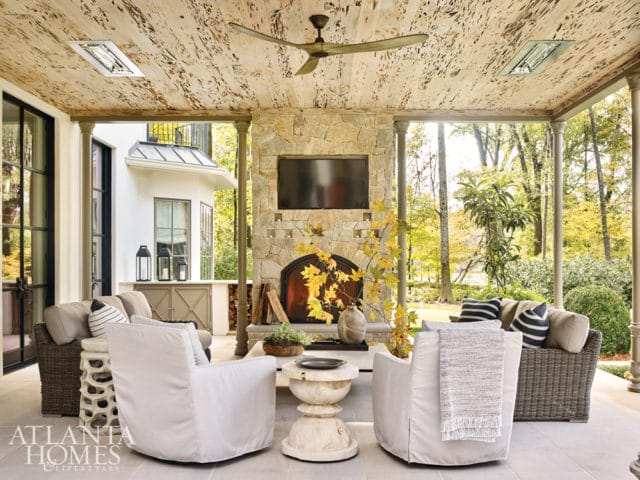 Covered patio with stone fireplace, TV, ceiling fan, and seating area with white slipcovered chairs and wicker sofa overlooking a wooded landscape.