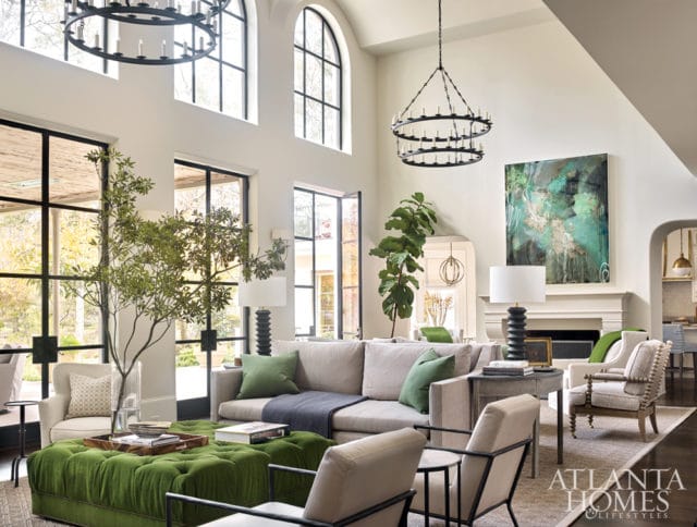 Living room with high ceilings, large arched windows, and two black chandeliers. Features a light gray sectional sofa, green tufted ottoman, and abstract green artwork above a fireplace.
