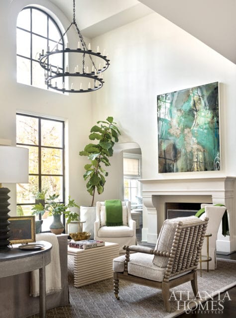 Living room with high ceilings, featuring a large arched window, a two-tiered black chandelier, a fireplace with abstract art above, and upholstered furniture including two armchairs and a sofa.