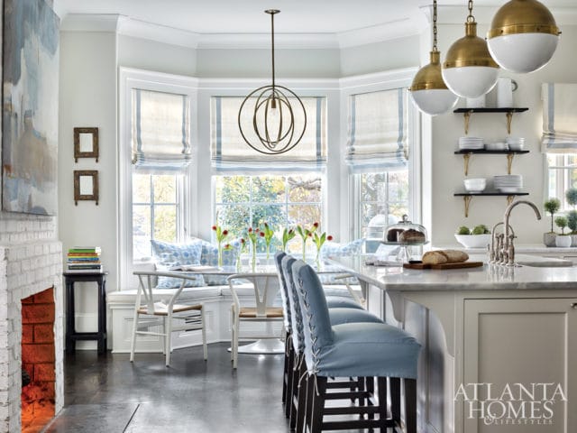Light-filled kitchen with white brick fireplace, marble island with blue upholstered bar stools, and bay window breakfast nook with striped roman shades.