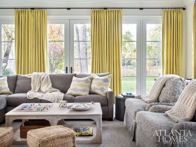 Gray sectional sofa and armchairs with patterned pillows and knit throws, in front of yellow curtains and French doors overlooking a garden and water.