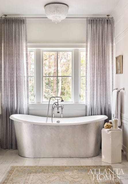 Freestanding bathtub with chrome fixtures in a bathroom with large windows, grey curtains, a crystal chandelier, and a patterned rug.