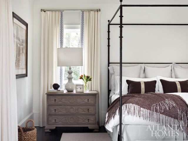 Bedroom with a black metal canopy bed, white linens, brown pillows, and a fringed throw. A bedside table with a lamp, books, and flowers sits next to a window with cream curtains.
