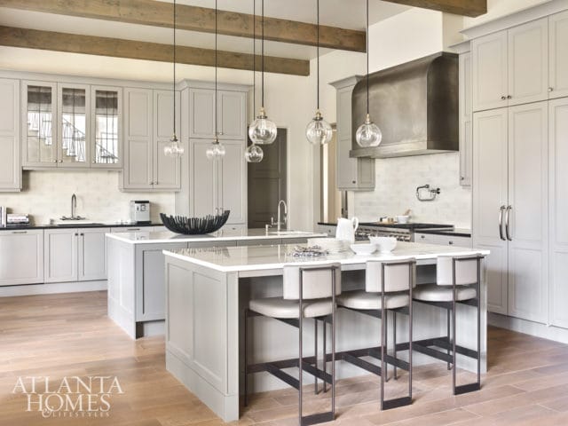 Gray kitchen with white subway tile backsplash, stainless steel range hood, and two islands with seating. Exposed wooden beams cross the ceiling.