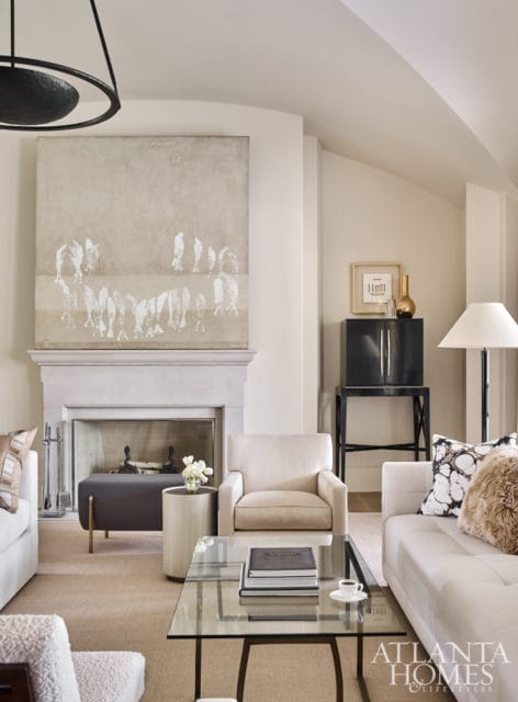 Living room with a large abstract painting above a stone fireplace, cream upholstered chairs, a glass coffee table, and a dark lacquered cabinet.