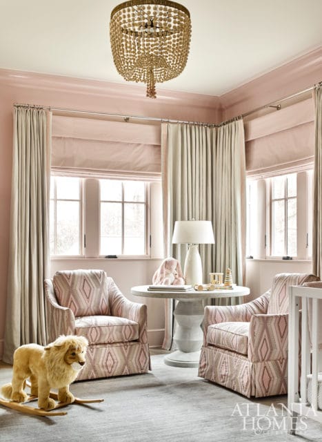 Nursery with pink walls, two upholstered armchairs with geometric pattern, round pedestal table with lamp and toys, and a rocking lion.