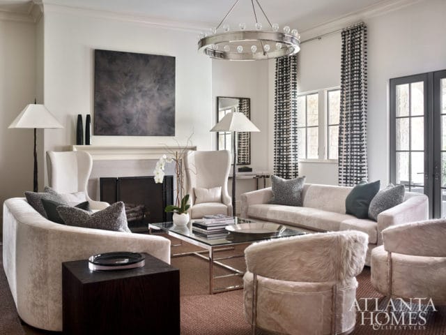 Living room with cream upholstered furniture, a glass and chrome coffee table, a fireplace with abstract art above, and black and white patterned curtains.