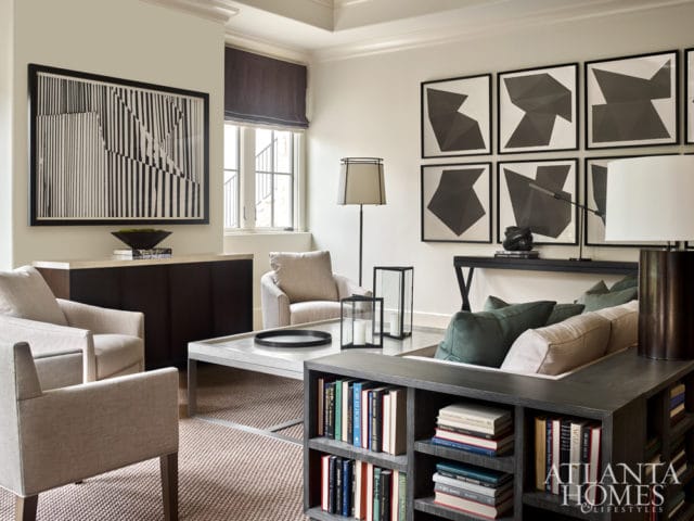 Living room with neutral upholstered chairs, a sectional sofa with green pillows, a white coffee table, and a dark wood bookshelf filled with books. Abstract art hangs on the walls.