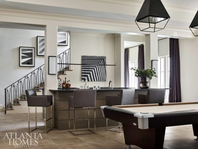 Modern bar area with dark wood cabinetry, leather bar stools, and a pool table in the foreground. Geometric pendant lights hang overhead.