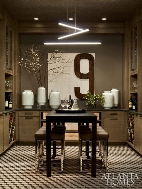 Modern wine cellar with wood cabinetry, glass-fronted cabinets, wine racks, a dark table with leather stools, and a large wooden number '9' art piece on the wall.