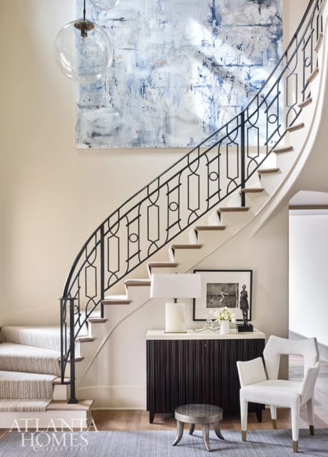 Curved staircase with black metal railing, abstract blue and white artwork, and a console table with a lamp, artwork, and sculpture.