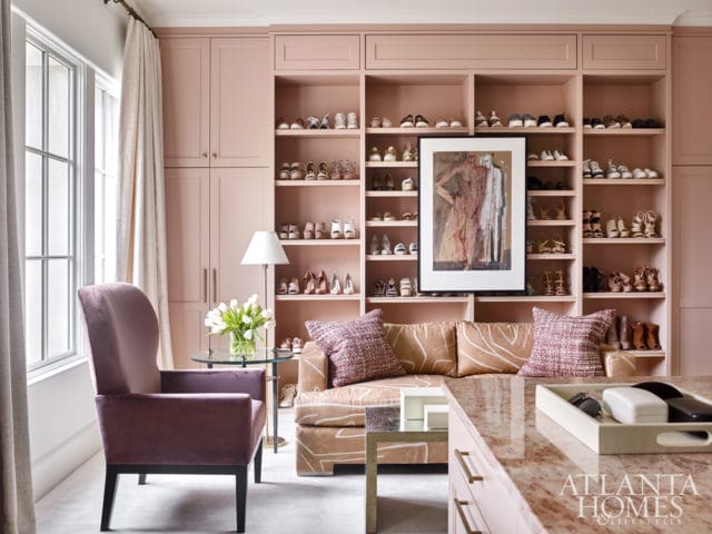 Pink built-in shelving unit filled with shoes, featuring a framed abstract artwork and a white lamp. A plush purple armchair and a patterned sofa are visible in the foreground.