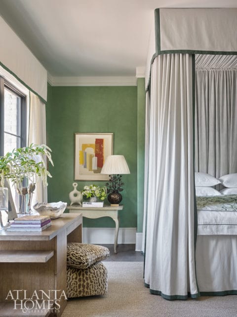 Green bedroom with a four-poster bed draped in white fabric, a wooden desk with leopard print ottomans, and abstract art on the wall.