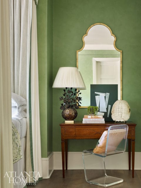 Green walls in a bedroom with a gold-framed mirror, a wooden console table with a lamp, books, and decorative objects, and a clear acrylic chair.