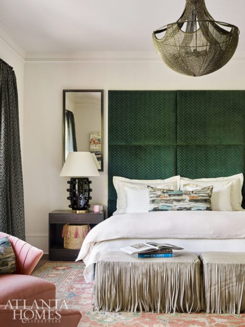 Bedroom with a large green upholstered headboard, white bedding, and a fringed ottoman. A patterned rug and a pink armchair are visible.