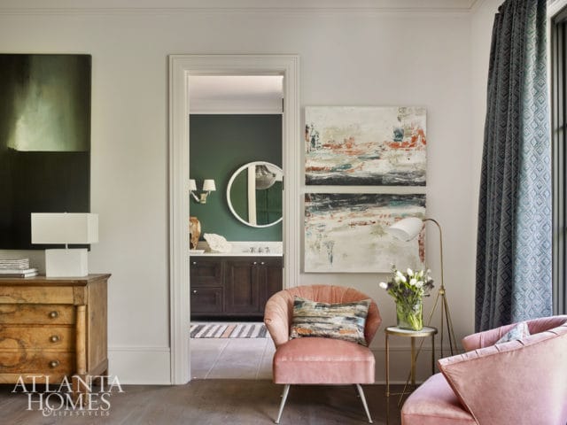 Living room with two plush pink armchairs, abstract art, and a doorway leading to a green bathroom with a round mirror and dark wood vanity.