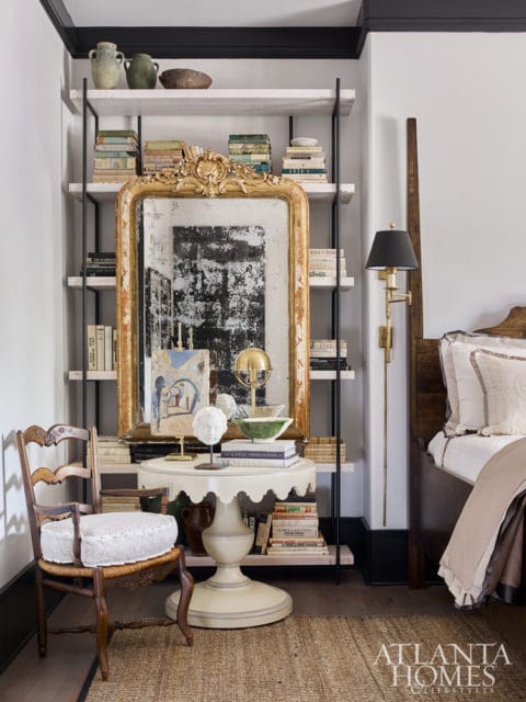 Bedroom with a tall wooden bed, a vintage chair, a round pedestal table with a gold ornate mirror, and bookshelves filled with books.