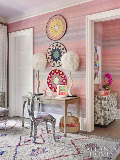 Pink striped wallpaper adorns a room with a light gray desk, ornate chair, and a fluffy white lamp. Three woven wall hangings hang above the desk.