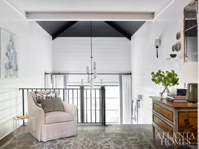 Loft living area with white shiplap walls, black vaulted ceiling, upholstered armchair with patterned pillows, and a vintage dresser with a mirrored wall sconce above.