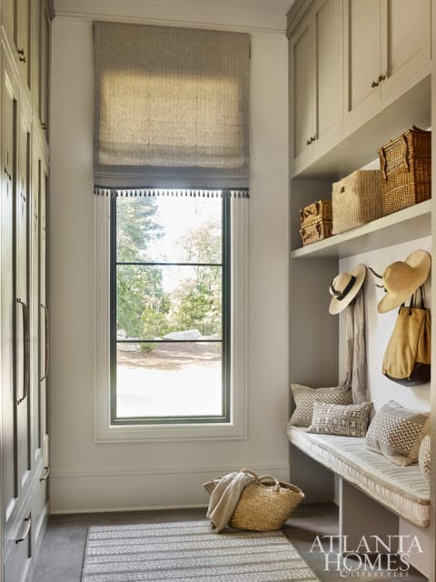 Mudroom with built-in gray cabinetry, a window with a linen Roman shade, a bench with patterned pillows, and woven storage baskets.