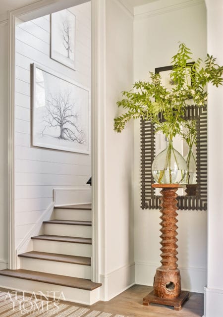 Hallway with shiplap walls, a staircase with dark wood treads, and a decorative wooden pedestal holding glass vases with greenery.
