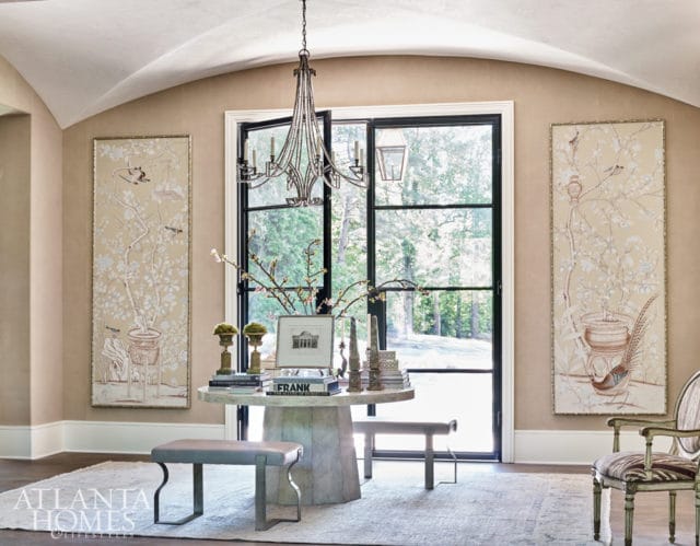 Foyer with a large round stone table, two benches, a crystal chandelier, and two tall framed artworks depicting birds and branches on beige walls.