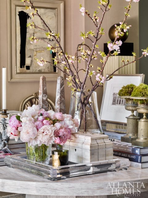 A table vignette featuring a vase of pink peonies, a decorative box, a framed artwork, and blooming branches on a marble table.