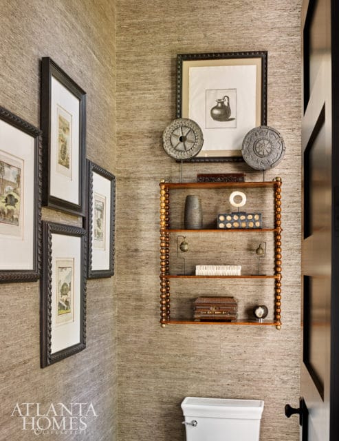 Powder room with textured taupe wallpaper, framed botanical prints, a wooden shelf unit with decorative objects, and a white toilet.