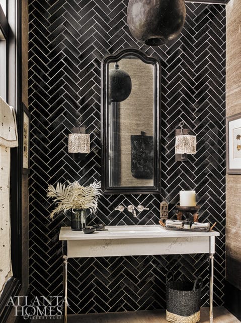 Powder room with black herringbone tile walls, white console sink with chrome faucets, ornate mirror, and decorative sconces.