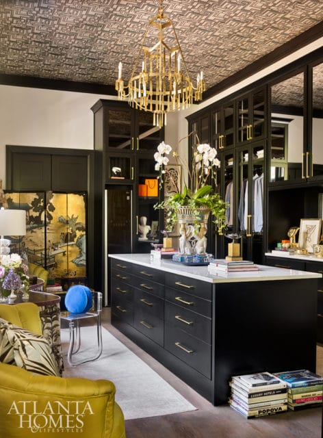 Black walk-in closet with a white marble-topped island, gold chandelier, patterned ceiling, and gold-accented cabinetry.
