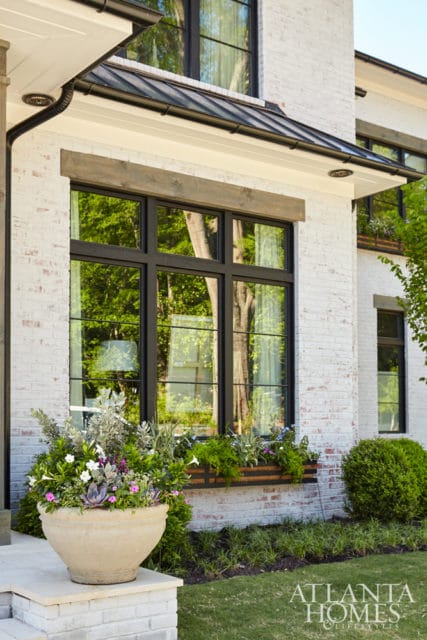 Modern home exterior with white painted brick, black-framed windows, a metal roof, and a large stone planter filled with greenery and flowers.