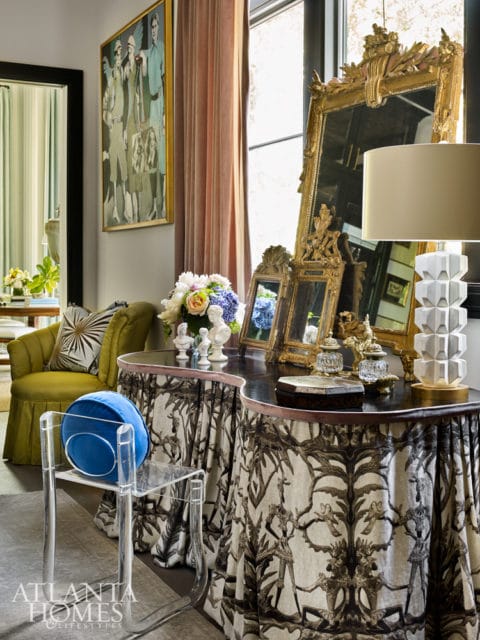Interior scene with a gold framed painting, velvet curtains, a green armchair, and a console table with ornate mirrors and a modern lamp.