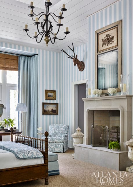 Bedroom with blue and white striped wallpaper, a dark wood bed with blue and white patterned bedding, a blue and white patterned armchair, and a stone fireplace with a deer head mounted above it.