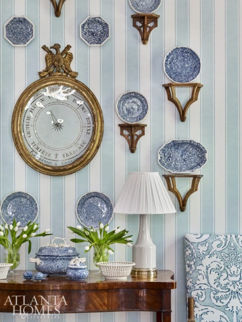 Blue and white striped wallpaper adorns a wall displaying a collection of blue and white plates, a gold-framed barometer, and a table with white tulips, blue ceramic dishes, a white lamp, and a chair with damask upholstery.