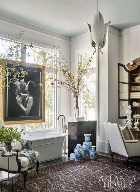 Freestanding bathtub in a bright bathroom with a black and white portrait, blue and white vases, and a patterned rug.