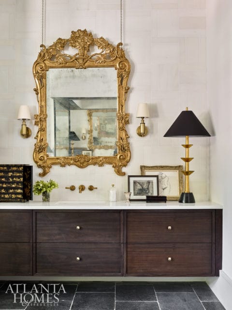 Bathroom vanity with dark wood drawers, white countertop, and ornate gold mirror above a sink. Brass sconces flank the mirror, and a black lamp sits on the counter.